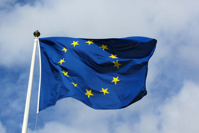 EU flag - blue background and circle of yellow stars - at top of white flag pole against cloudy blue sky 