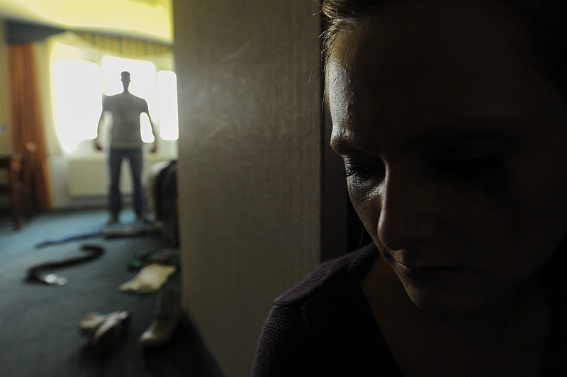 Image of a man standing in a room in disarray and a women hiding behind the door.