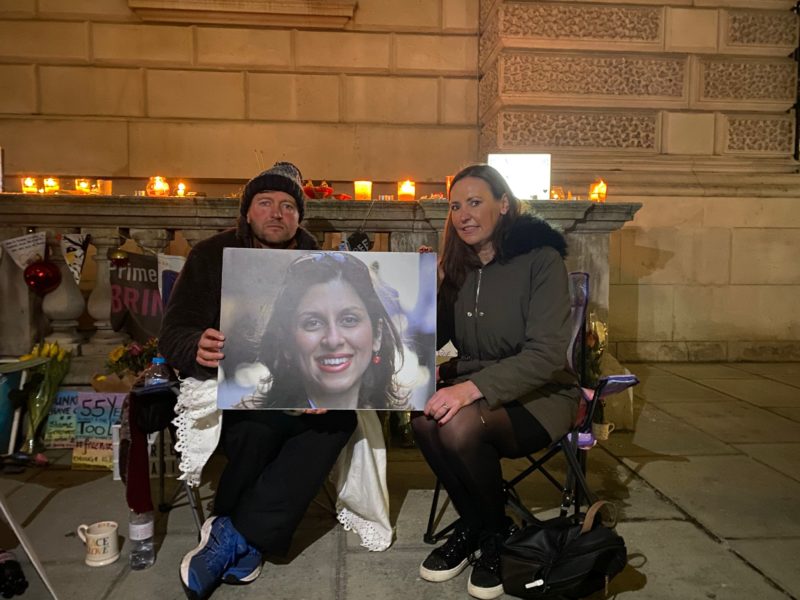 Photo of Vicky visiting Richard Ratcliffe outside the FCDO. They are sitting in camping chairs and holding a photo of Nazanin between them, with lit candles, flowers and placards in the background.