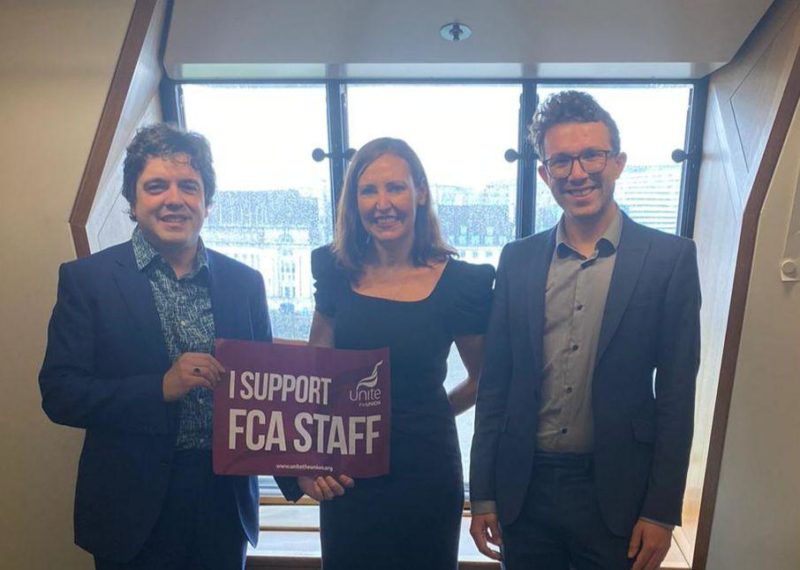 Two FCA Staff (one on the left and one on the right) with Vicky Foxcroft (in the middle) standing by the window holding a poster saying " I support FCA Staff"