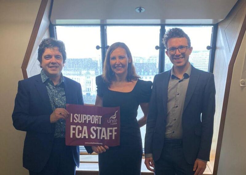 Two FCA Staff (one on the left and one on the right) with Vicky Foxcroft (in the middle) standing by the window holding a poster saying " I support FCA Staff"