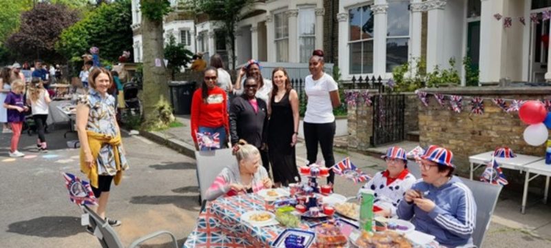 Photo of Vicky standing with five residents of Kitto Road at the Jubliee Street Party. Three other residents are seated at a Union Jack-covered table in front of them enjoying some party food.